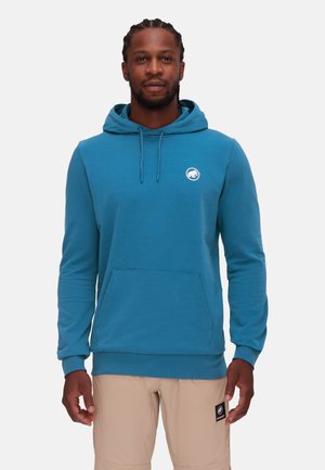 Mann trägt blauen Hoodie mit Fronttasche und beigefarbene Shorts, steht vor einem schlichten hellen Hintergrund.