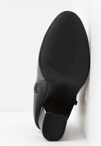 Zapato de cuero negro con puntera redondeada y tacón bajo cuadrado; presenta una suela texturizada y un pequeño detalle de costura decorativa.