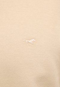 Beige fabric close-up with small embroidered horse logo in matching beige thread.