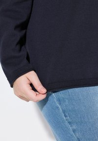 Dunkelblaues Strickpullover mit gerippten Bündchen und Saum, kombiniert mit hellblauen Jeans. Hand greift den Saum, um die Textur und Passform zu zeigen.