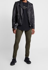 Černá kožená motocyklová bunda s texturovaným povrchem, spárovaná s černým tričkem a úzkými kalhotami olivové barvy. Celkový vzhled doplňují černé tenisky.