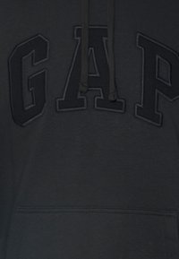 Black hoodie featuring an embroidered "GAP" logo in a slightly lighter shade, with a front pocket and drawstring hood. Soft fabric texture.