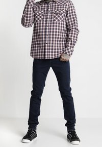 Camicia a maniche lunghe a quadri rossa, blu e bianca; jeans scuri; sneaker nere con suola bianca; mani tatuate visibili.
