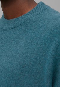 Tyrkysový pletený svetr s kulatým výstřihem. Měkká textura a volný střih, ukazující hladký a rovnoměrný povrch látky.