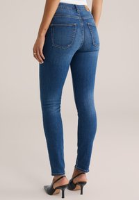 Modré džíny skinny mají vysoký pas, dvě zadní kapsy a čistý vzhled. Jsou kombinovány s černými botami na vysokém podpatku.