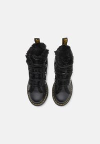 Pár černých kožených šněrovacích bot s černým kožíškem a žlutým prošíváním kolem podrážky, z pohledu shora na bílém pozadí.