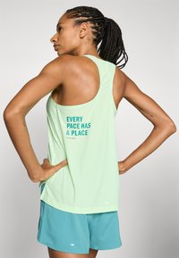 Top deportivo sin mangas de color verde claro con textura de malla. Presenta texto en la parte de atrás: "CADA RITMO TIENE SU LUGAR" en color teal. Combinado con pantalones cortos azules.