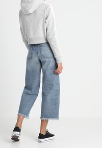 Hoodie grigia corta con cappuccio, abbinata a jeans larghi blu chiaro con orli sfrangiati. Sneakers nere completano l'outfit.