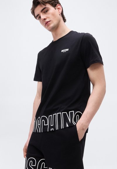 Νεαρός άνδρας που φοράει μαύρο t-shirt Moschino Active και αντίστοιχα σορτς με έντονα λευκά γράμματα, στέκεται με το ένα χέρι στην τσέπη.