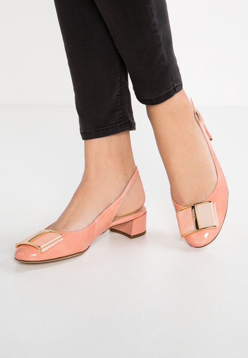 Pieds portant des chaussures slingback roses à talon bas avec des détails de nœud en or et tissu, associés à un pantalon noir court sur fond blanc.