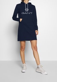 Marineblauwe hoodie-jurk met een kangoeroezak, witte koordjes en een groot GANT-logo op de borst. Heeft lange mouwen en een relaxte pasvorm.