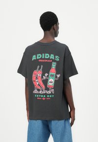T-shirt nera in cotone con una stampa grafica colorata che raffigura peperoncini e una bottiglia di salsa piccante, etichettata "ADIDAS ORIGINALS EXTRA HOT."