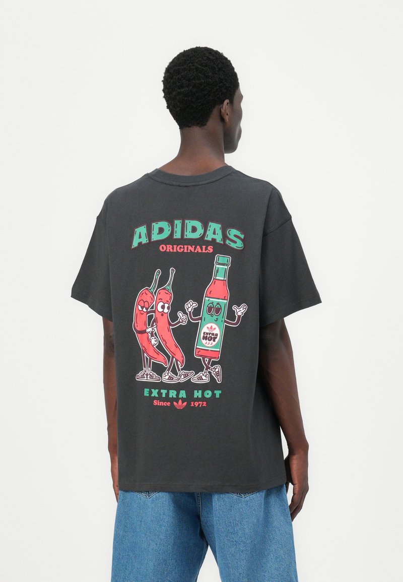 T-shirt nera in cotone con una stampa grafica colorata che raffigura peperoncini e una bottiglia di salsa piccante, etichettata "ADIDAS ORIGINALS EXTRA HOT."