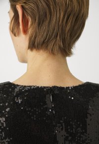 Svart paljettkjole med teksturert stoff, har rund hals og glidelås bak. Paljettene reflekterer lyset, noe som skaper en glitrende effekt.