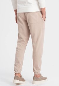 Beige, tapered Jogginghosen aus weichem Stoff mit elastischen Bündchen und Seitentaschen, kombiniert mit grauen Sneakers auf einem weißen Hintergrund.