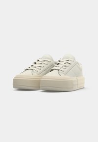 Sneakers low-top di colore bianco sporco con suole a piattaforma spesse e testurizzate e lacci abbinati di colore bianco sporco su uno sfondo semplice.