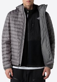 Grå puffjacka med kviltat mönster, huva och dragkedja framtill. Lagd över en svart fleece- och nylonkombination. Texturerade tygdetaljer.