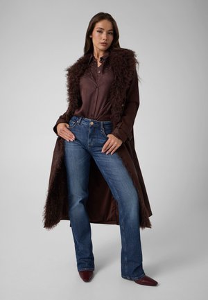 Femme debout portant un manteau marron foncé doublé de fourrure par-dessus une chemise marron, un jean bleu et des chaussures sombres pointues sur un fond gris uni.