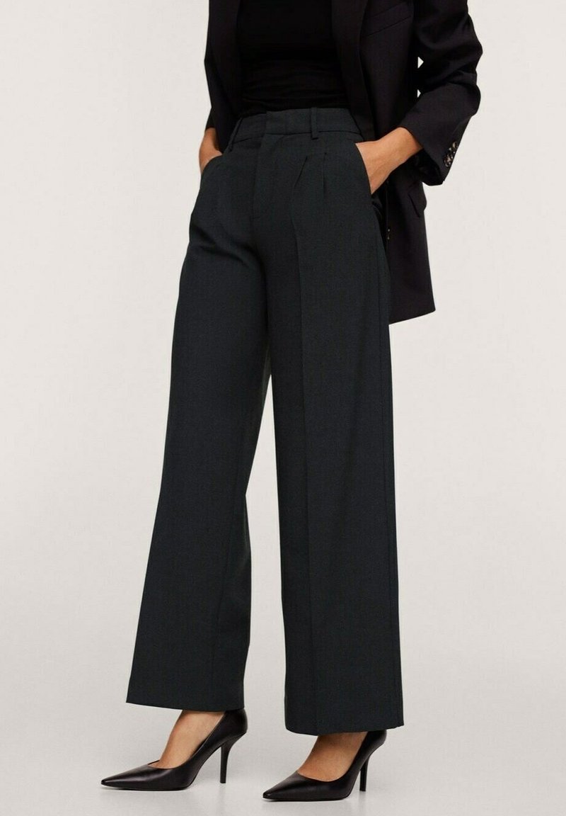 Pantalons larges taille haute de couleur vert foncé en tissu texturé, avec des plis sur le devant et des poches latérales. Associés à des talons noirs.