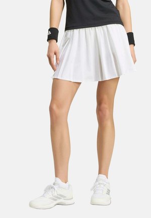 Persona con falda blanca plisada, camisa negra, muñequeras negras, zapatillas deportivas blancas, de pie con las piernas ligeramente separadas.