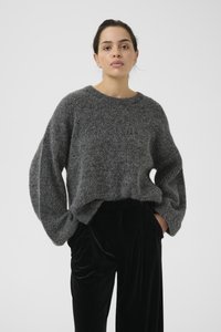 Dik grijs gebreide trui met oversized mouwen, gek textureerd oppervlak, ronde halslijn, gedragen over zwarte fluwelen broeken met plooien.