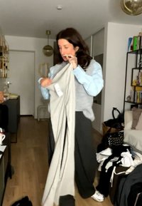 Frau faltet hellgraue Hose in einem Wohnzimmer mit Holzboden, Bücherregalen und verstreuter Kleidung auf einem Stuhl und am Boden.