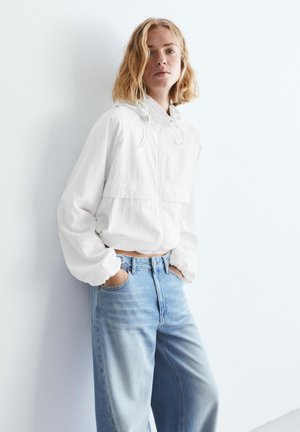 Młoda kobieta ubrana w białą bluzę z kapturem i zamkiem oraz jasnoniebieskie dżinsy, stojąca z rękami w kieszeniach na tle białej ściany.