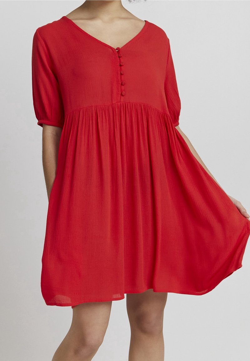 Femme portant une robe rouge ample avec manches aux coudes, encolure en V boutonnée et taille froncée, tenant légèrement la jupe sur le côté.