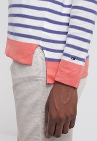 Zebrowany sweter w kolorze białym i granatowym z koralowymi akcentami. Posiada ribbowany dół, boczne rozcięcia i naszywkę z logo na rękawie. Wykonany z lekkiego materiału.