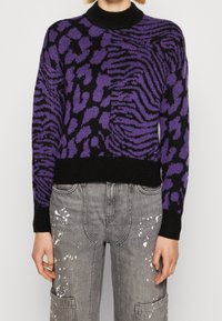 Pull imprimé animal violet et noir avec un col noir côtelé. Le tissu semble tricoté et présente une coupe décontractée, associé à un pantalon gris éclaboussé de peinture.