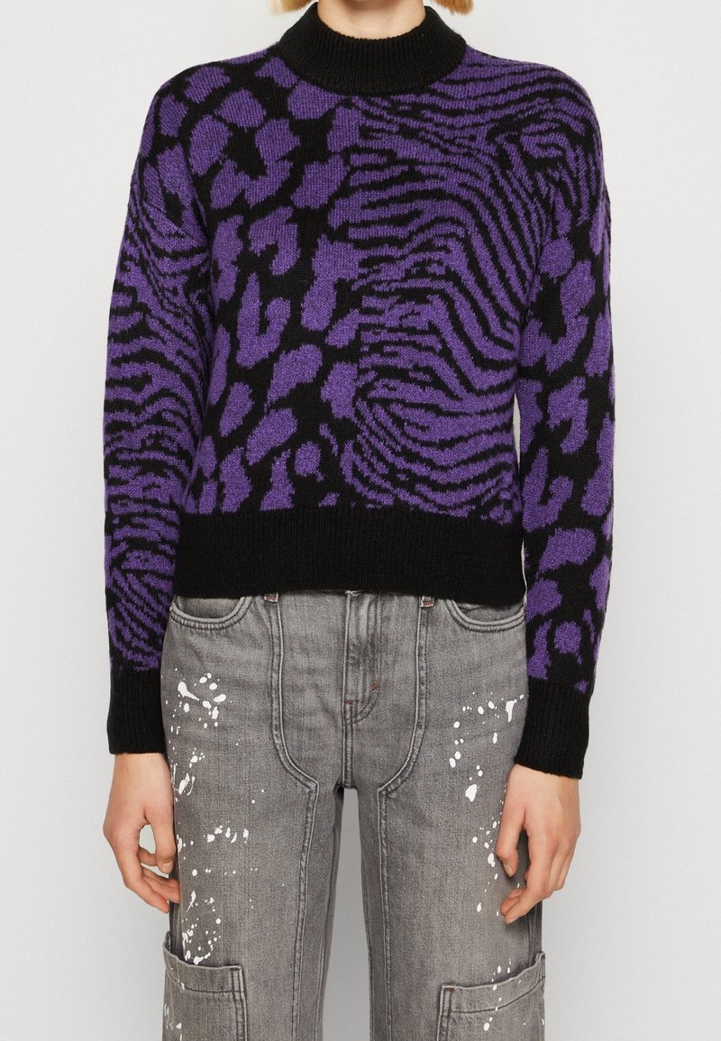 Pull imprimé animal violet et noir avec un col noir côtelé. Le tissu semble tricoté et présente une coupe décontractée, associé à un pantalon gris éclaboussé de peinture.