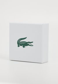 Boîte rectangulaire blanche avec une surface texturée, arborant un logo de crocodile vert à l'avant. Design simple sans motifs supplémentaires.