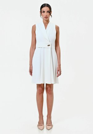 Femme aux cheveux foncés portant une robe blanche sans manches avec un bouton doré, debout sur un fond blanc uni, portant des escarpins pointus couleur chair.