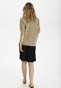 Donna con lunghi capelli mossi, che indossa un maglione leggero a maglia e una gonna nera, in piedi di spalle rispetto alla macchina fotografica su uno sfondo neutro.