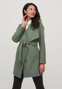 Abrigo verde envuelto de tela texturizada, con cinturón en la cintura, mangas largas y un diseño de cuello abierto.