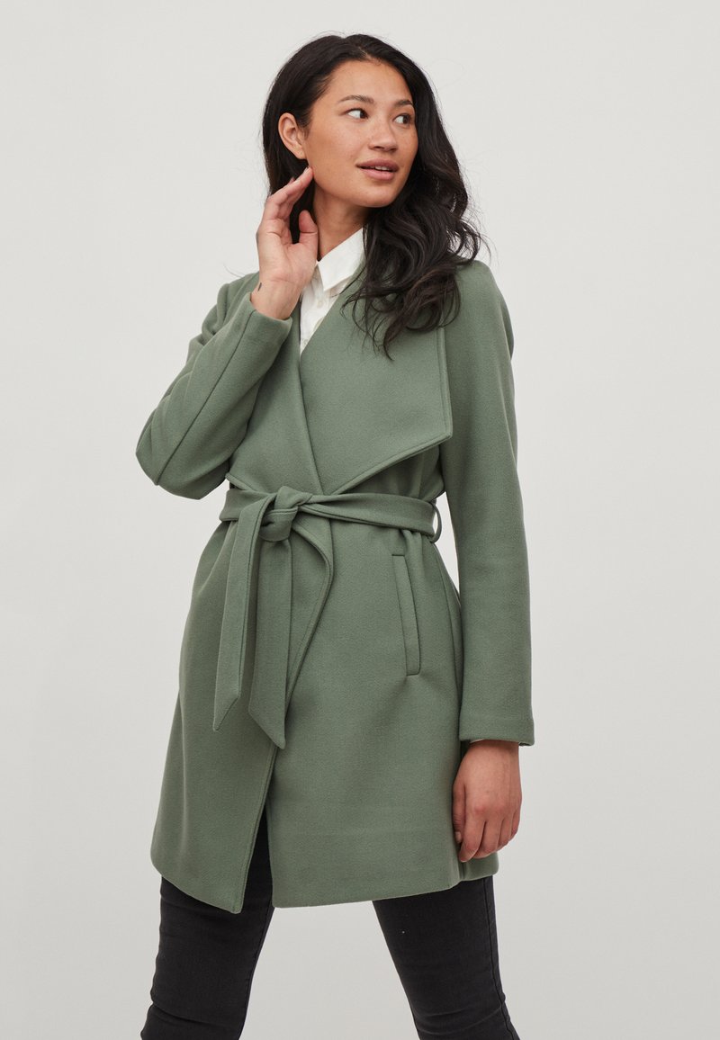 Abrigo verde envuelto de tela texturizada, con cinturón en la cintura, mangas largas y un diseño de cuello abierto.