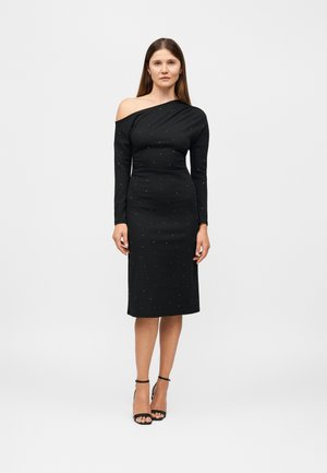 Une femme porte une robe midi noire ajustée, ornée de clous discrets, avec une encolure asymétrique à une épaule, ainsi que des chaussures noires à talons et bride cheville.