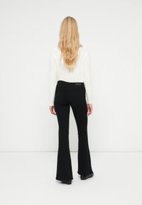 Nainen, jolla on pitkät vaaleat hiukset, pukeutuneena valkoiseen neuleeseen ja mustiin leveälahkeisiin housuihin, seisoo valkoista seinää vasten, kädet rentoina sivuilla.