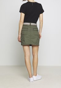 Oliwkowa spódnica z dwiema tylnymi kieszeniami, elastycznym pasem i wzorzystym paskiem. W zestawie z gładką czarną koszulką i białymi sneakersami.