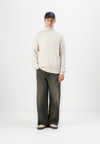 Young man wearing a cream turtleneck sweater, black cap, wide-leg faded black jeans, and white sneakers standing against a plain white background.