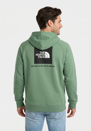 Uomo che indossa una felpa verde con cappuccio, logo quadrato nero e testo "THE NORTH FACE" e "NEVER STOP EXPLORING", visto da dietro su sfondo bianco.