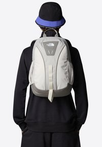Zaino grigio e beige con spallacci imbottiti, tasche laterali in rete e un logo sul davanti. Tessuto liscio e resistente con una forma strutturata.