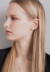 Auksinės spalvos kaklo grandinėlė su širdies formos pakabuku, dėvima žmogaus su ilgu tiesiu šviesiai auksiniu plaukų ant šviesiai pilko fono.