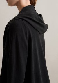Schwarze Kapuzenjacke aus weichem Stoff, mit lockerer Passform und einer Kapuze auf der Rückseite. Glatte Textur mit minimalistischem Design.