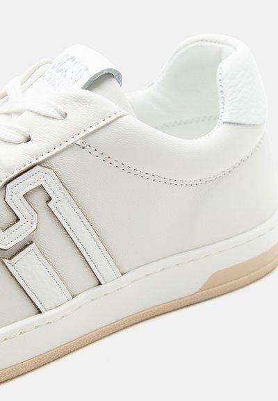 Baskets en cuir blanc avec lacets blancs, détails de design surpiqués, et semelle en caoutchouc beige, montrées en vue rapprochée de côté.