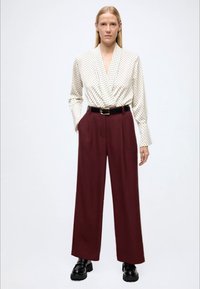 White blouse with black dots, deep burgundy wide-leg trousers, black belt, and chunky black shoes; smooth fabric and tailored design.