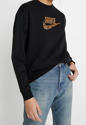 Persoon met een zwart sweatshirt met een Nike-logo in luipaardprint en lichtblauwe spijkerbroek, hand rustend bij de zak.