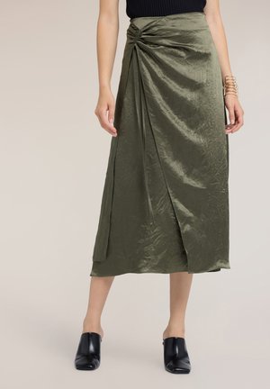 Kvinde iført en skinnende olivengrøn omslagsnederdel med rynket detalje ved taljen, sorte slip-on sko og en sort ærmeløs top.