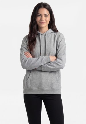 Jonge vrouw met lang donker haar draagt een grijze hoodie en zwarte broek, staat met de armen gekruist en glimlacht zachtjes tegen een egale achtergrond.