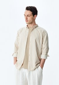 Camisa de botones beige a rayas con mangas arremangadas sobre un top de punto beige, combinada con pantalones de lino de color claro. Diseño simple, textura suave.